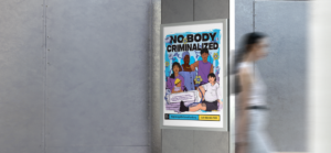 Person walks in front of a poster showing five people and the title "No Body Criminalized"