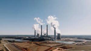 A wide view of a coal-fired power plant with multiple tall smokestacks emitting white smoke into a clear blue sky. The plant is surrounded by open fields, a water reservoir, and industrial structures, emphasizing the industrial and environmental impact of coal energy production.